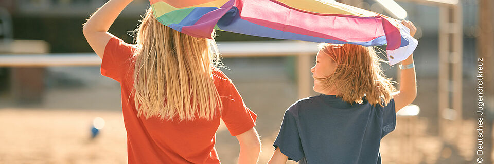 Zwei Kinder sind von hinten zu sehen. Sie laufen bei Sonnenschein auf einen Spielplatz und halten gemeinsam eine Regenbogenfahne über ihre Köpfe.