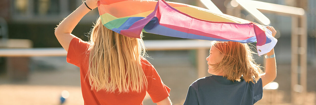 Zwei Kinder sind von hinten zu sehen. Sie laufen bei Sonnenschein auf einen Spielplatz und halten gemeinsam eine Regenbogenfahne über ihre Köpfe.